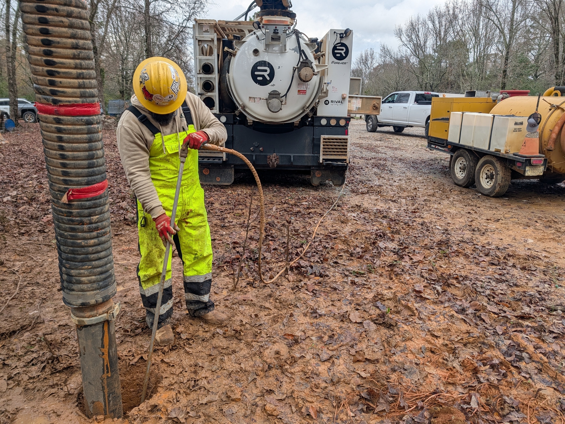 Hydrovac Excavation - A Better Way to Dig A worker in a yellow safety suit and hard hat uses a hose to vacuum mud from a hole at a construction site, with heavy equipment and trucks in the background on a cloudy day.