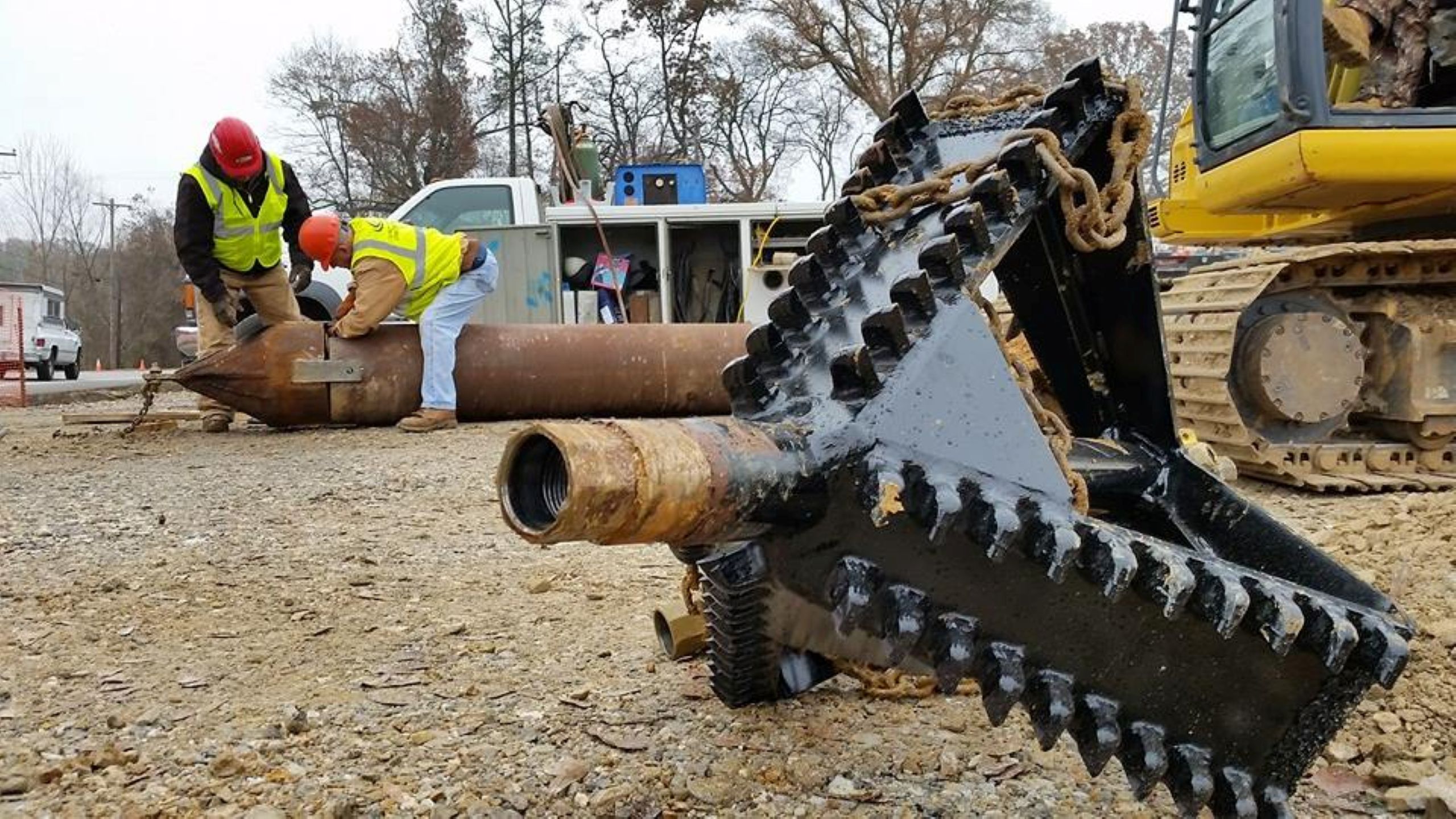 Two workers in safety gear work near a large metal pipe on a construction site. In the foreground is a close-up of a heavy, jagged industrial tool lying on the ground. Construction vehicles and trees are in the background.