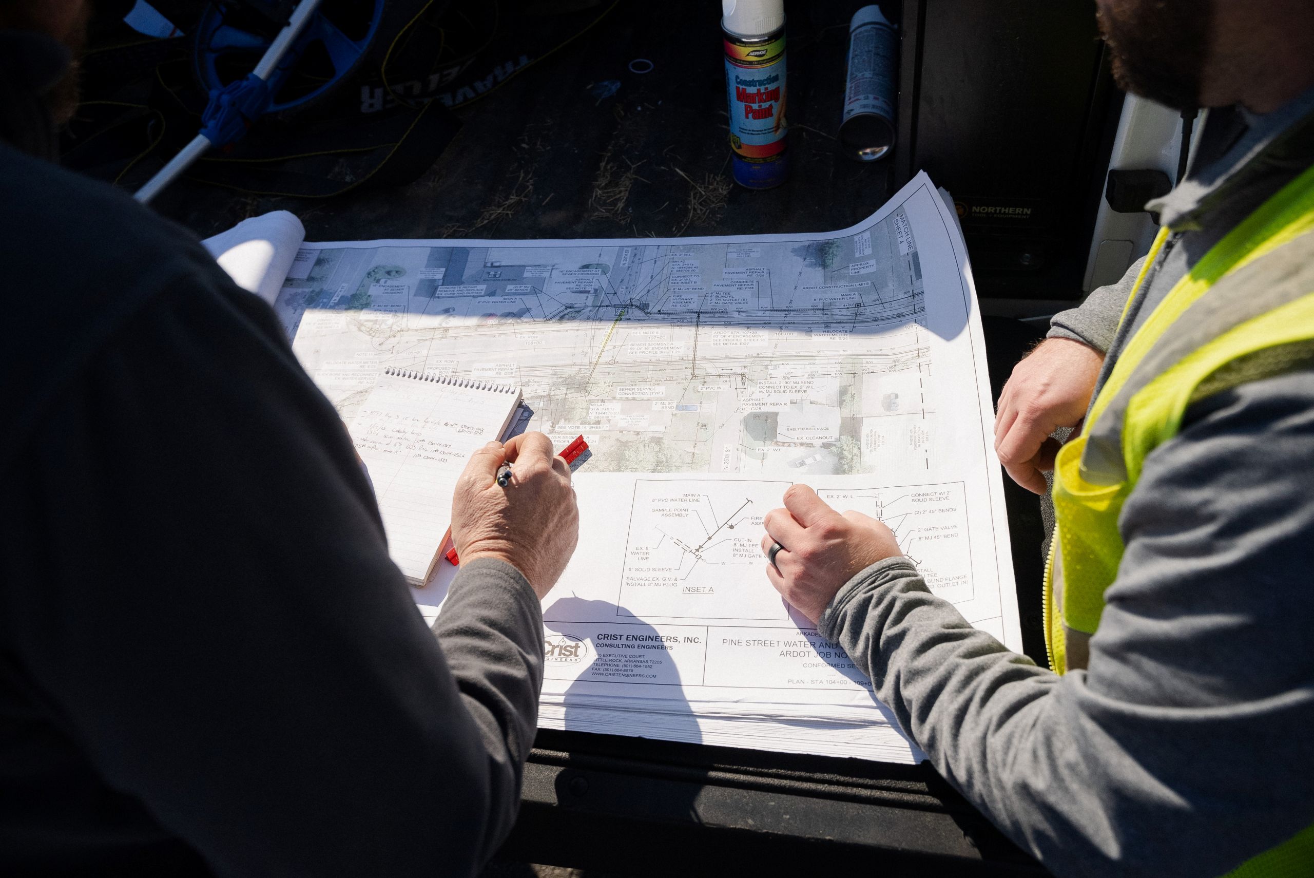 Two people review a large architectural blueprint outdoors; one holds a red marker and the other wears a safety vest, indicating a construction or planning discussion.