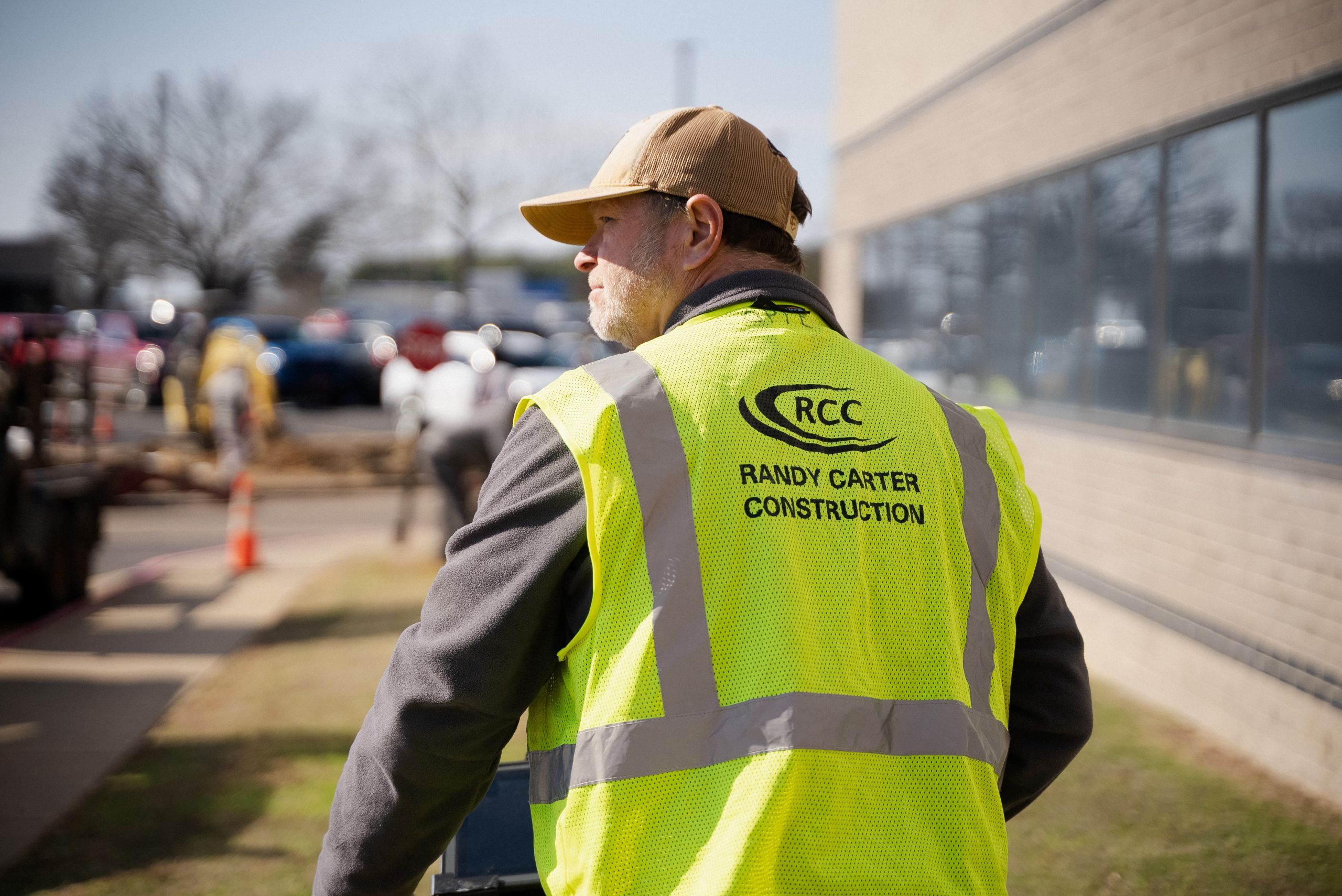 A man in a yellow Randy Carter Construction safety vest and tan cap stands outside near a building, looking to the side. Construction activity and parked cars are visible in the background.
