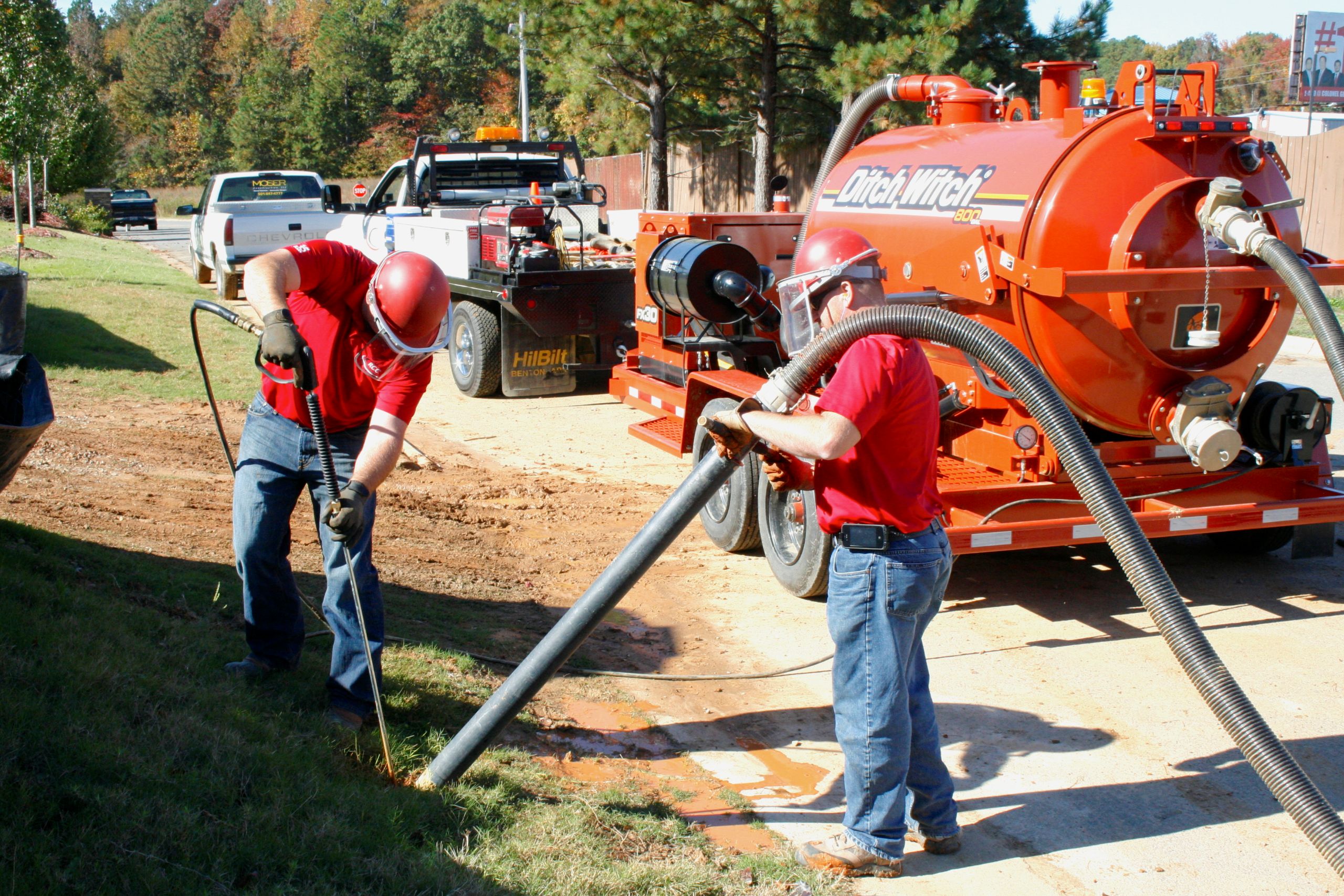 Two workers in red shirts and helmets use hoses connected to a large orange vacuum truck, performing excavation work on a grassy roadside near other parked utility trucks.